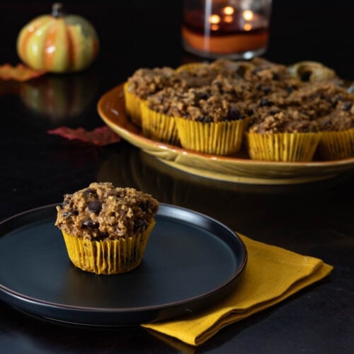 Pumpkin Pecan Muffin sitting on a black plate with a large pumpkin shaped serving plate in the background with the remaining muffins, decorated with a fall theme.