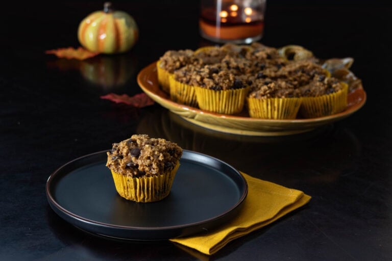 Pumpkin Pecan Muffin sitting on a black plate with a large pumpkin shaped serving plate in the background with the remaining muffins, decorated with a fall theme.