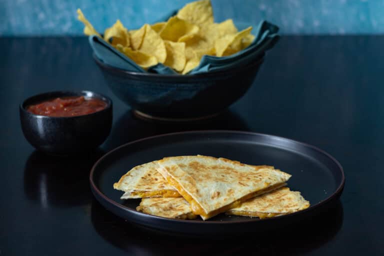Quesadilla divided into four pieces sitting on a black plate with a big bowl of chips and salsa sitting in the background.
