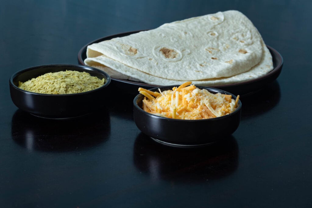 High Protein Quesadilla ingredients sitting on a table - nutritional yeast, vegan shredded cheese, and tortillas.