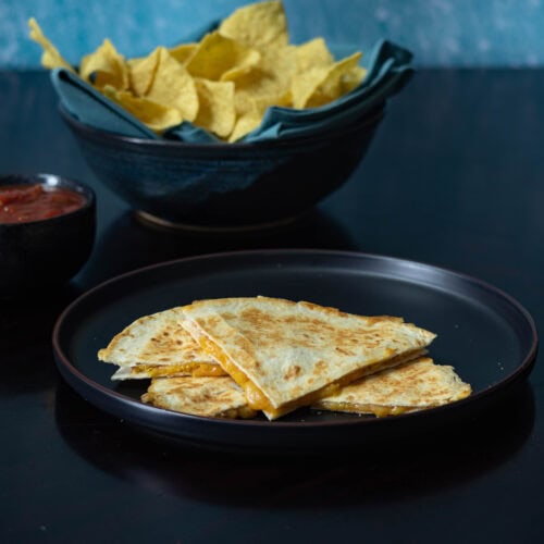 Quesadilla divided into four pieces sitting on a black plate with a big bowl of chips and salsa sitting in the background.