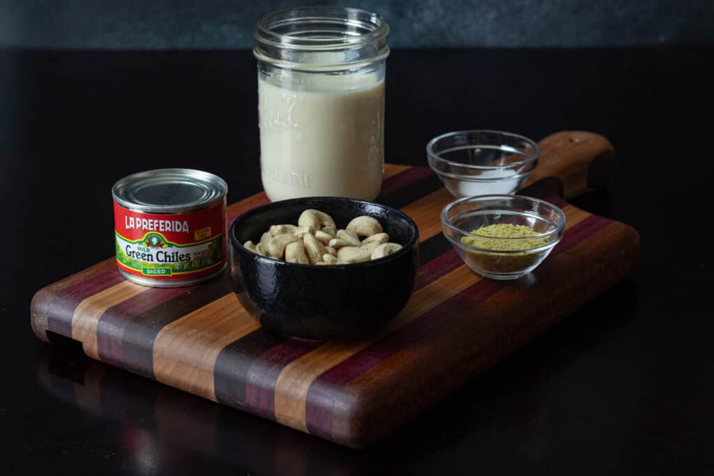 Homemade vegan queso ingredients sitting on a wooden cutting board - can of green chilies, raw cashews, soy milk, salt and nutritional yeast.