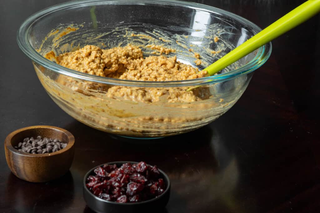 Muffin batter in large glass bowl, next to small bowls filled with chocolate chips and craisins.