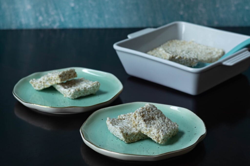 Two small plates with two rice crispies each stacked, with a small square baking dish in the background.