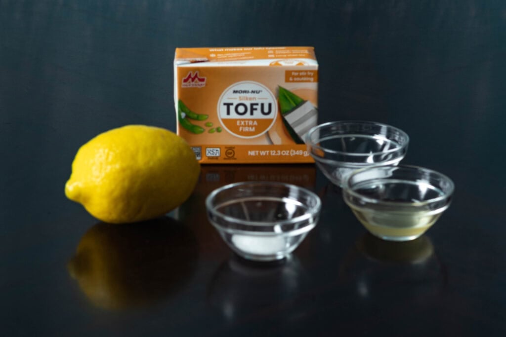 High protein vegan sour cream ingredients sitting on a table - lemon, silken tofu, salt, sugar, and vinegar.