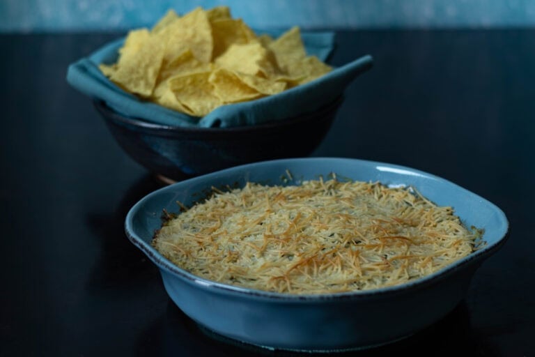 Round baking dish with artichoke dip and bowl of tortilla chips in the background.