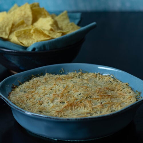 Round baking dish with artichoke dip and bowl of tortilla chips in the background.