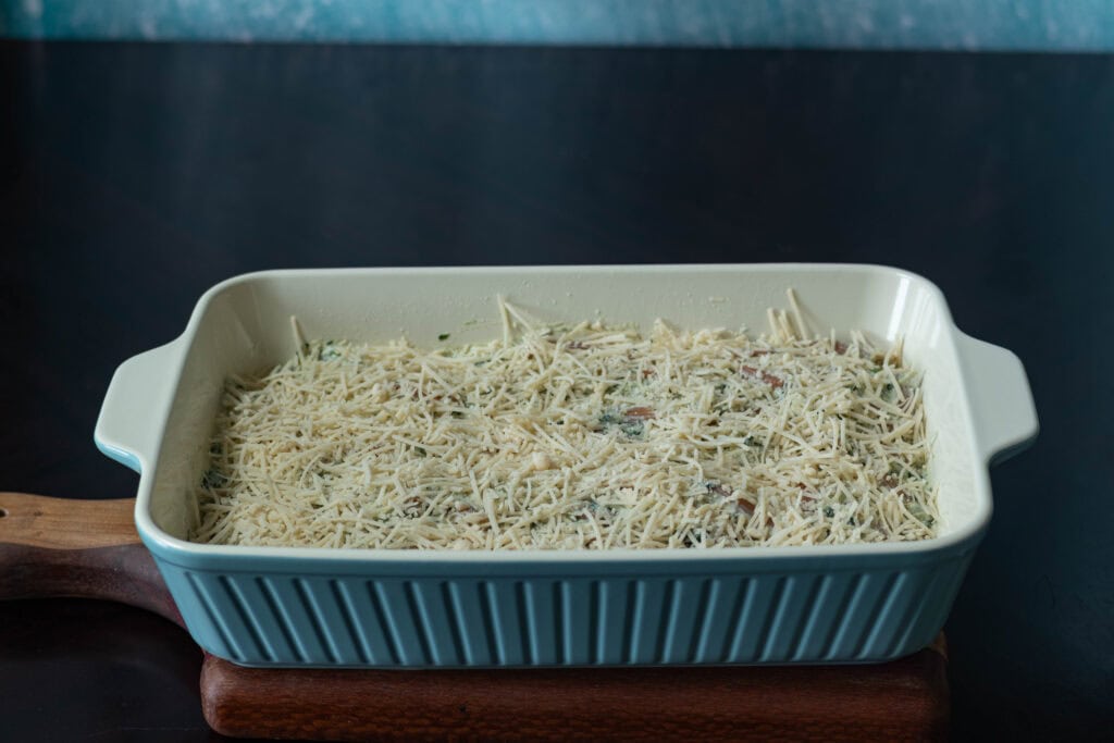 Large casserole dish with uncooked spinach and artichoke pasta topped with vegan parmesan.