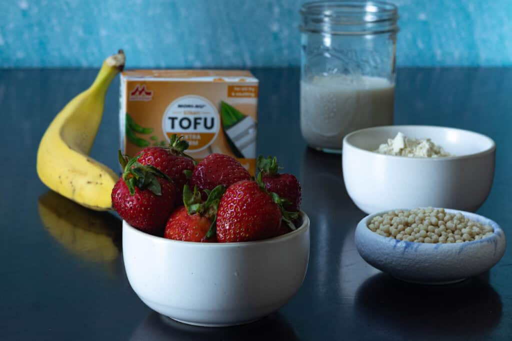 Strawberry banana smoothie bowl ingredients sitting on a table - banana, strawberries, silken tofu, soy milk, protein powder and edamame puffs.