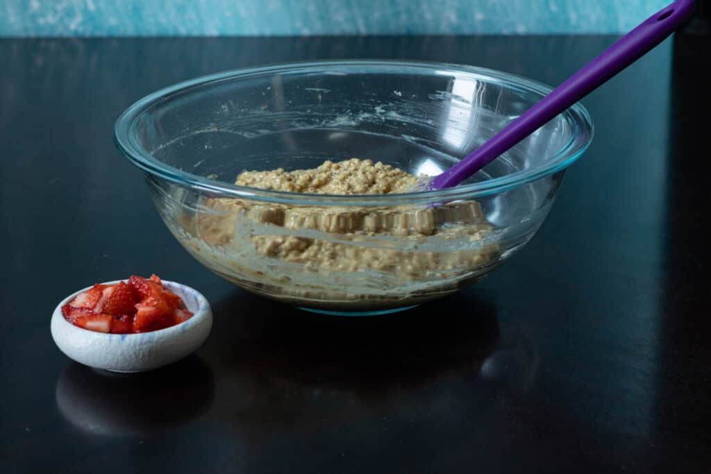 Large glass bowl with muffin batter sitting next to a smaller bowl of small strawberries chunks.
