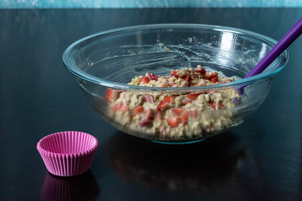 Strawberry muffin batter in large glass bowl sitting next to muffin liners.