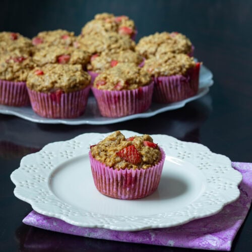 Small white flower-shaped plate with a strawberry muffin, large plate with remaining strawberry muffins in the background.