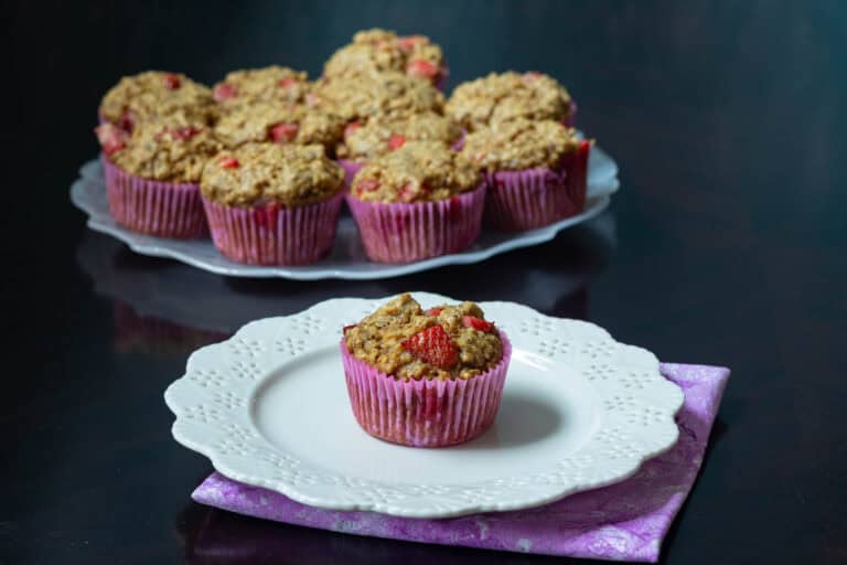 Small white flower-shaped plate with a strawberry muffin, large plate with remaining strawberry muffins in the background.