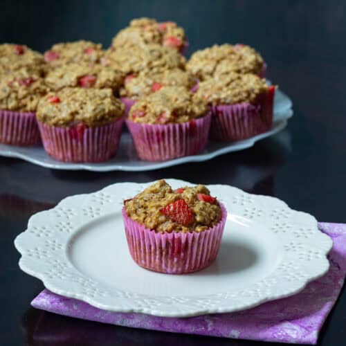 Small white flower-shaped plate with a strawberry muffin, large plate with remaining strawberry muffins in the background.