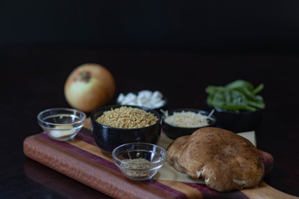 Ingredients for Stuffed Portobellos sitting on a wooden cutting board - large portobello mushroom caps, ground flax seed, TVP, seasoning, onion, vegan feta, vegan parmesan, and baby spinach.