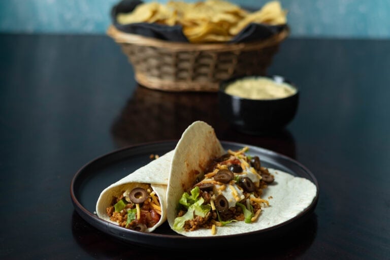 Large plate with two TVP tacos, and a basket of tortilla chips and cheese sauce in the background.