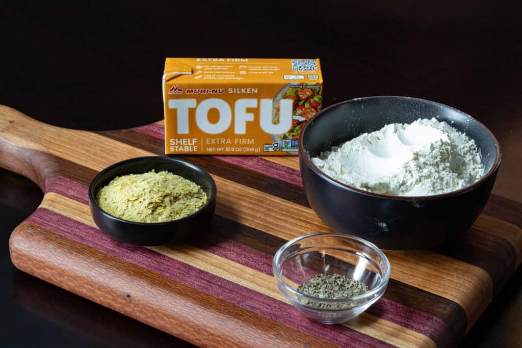 Pizza crust ingredients sitting on a wooden cutting board - spices, nutritional yeast, silken tofu and flour.
