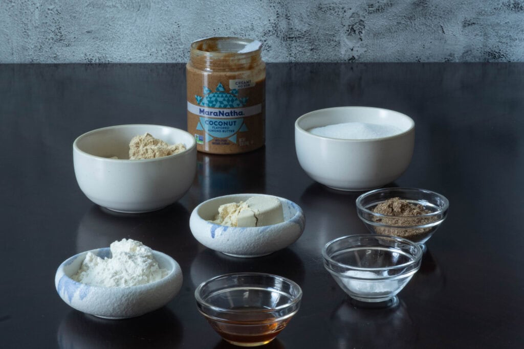 Almond butter cookie ingredients sitting on a wooden table - almond butter, sugar, almond protein powder, vanilla protein powder, flour, vanilla extract, coconut extract, ground flax seed, salt, baking powder and baking soda.