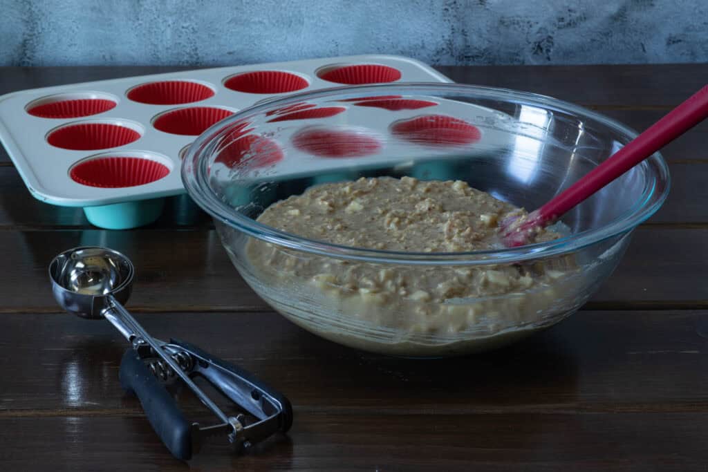 Apple Cinnamon Muffin batter in a large glass bowl, sitting next to an ice cream scooper and lined muffin tin.