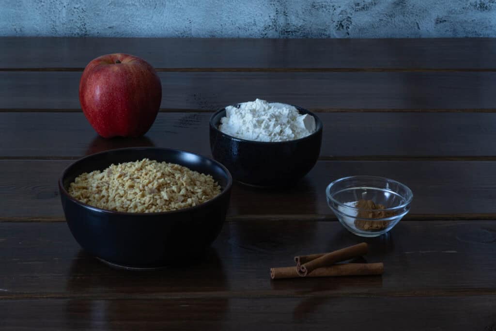 Ingredients for apple cinnamon muffins sitting on a wooden table - TVP, flour, an apple, cinnamon, salt, baking soda, baking powder and pumpkin pie spice.