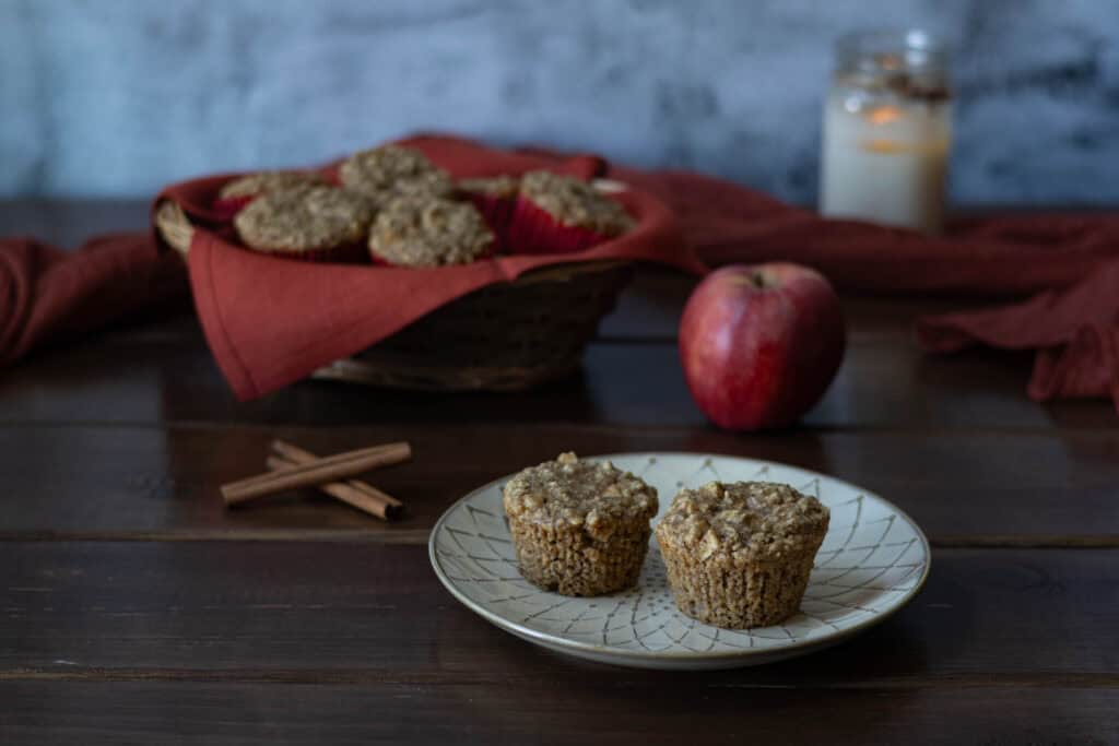 Two apple cinnamon muffins sitting on a textured plate, with an apple, cinnamon sticks, lit candle and basket of muffins in the background.