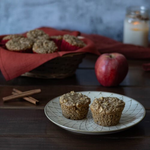 Two apple cinnamon muffins sitting on a plate, with a basket of muffins in the background, as well as an apple, candle and cinnamon sticks.