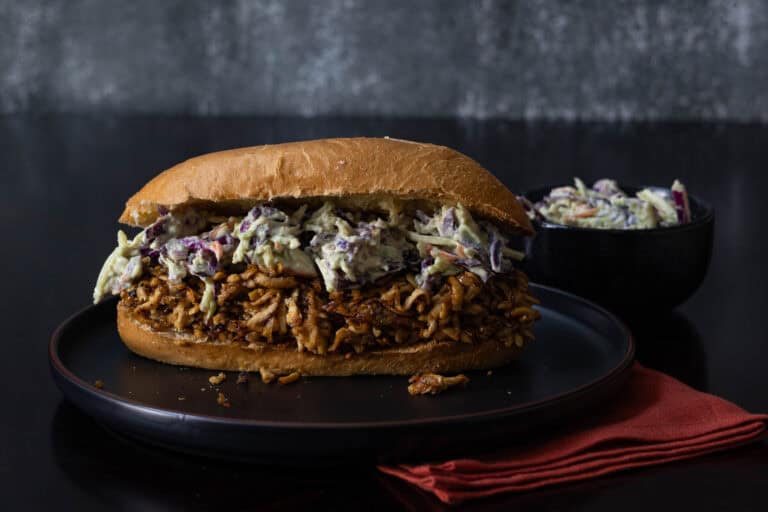 Shredded BBQ Tofu Coleslaw Sandwich on a toasted bun with a cup of cole slaw in the background.