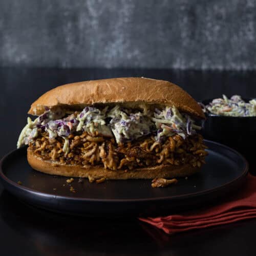 Shredded BBQ Tofu Coleslaw Sandwich on a toasted bun with a cup of cole slaw in the background.
