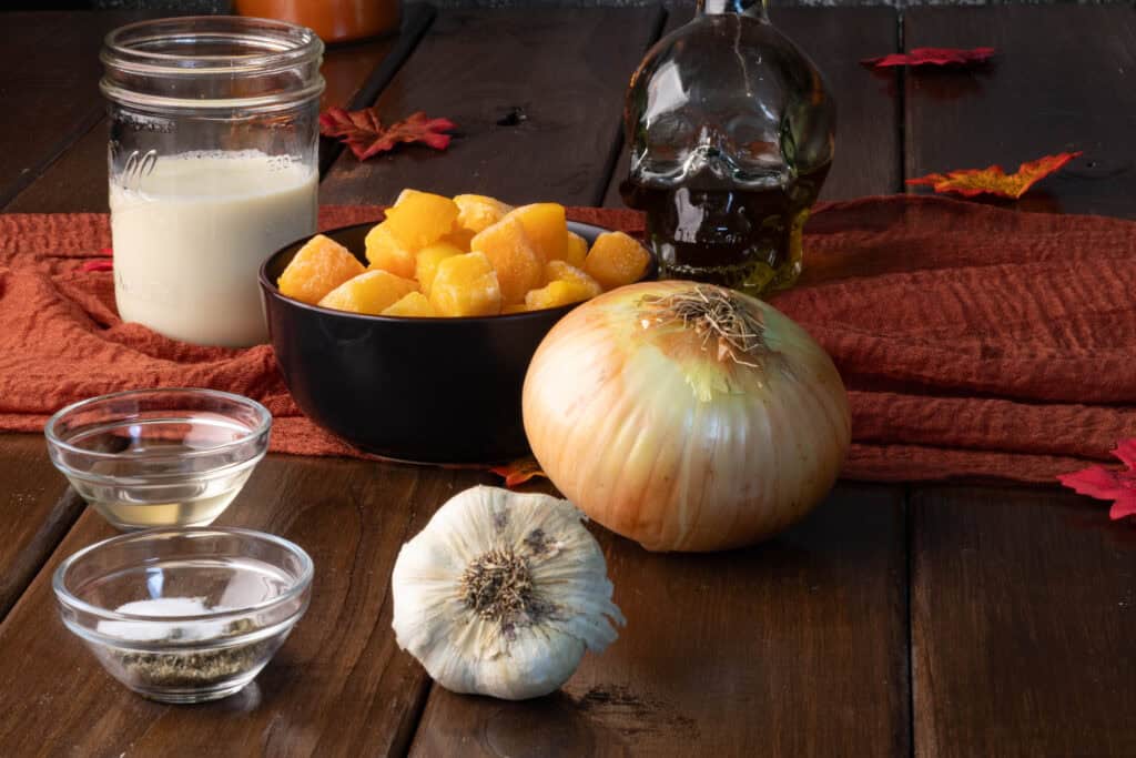 Butternut squash sauce ingredients sitting on a wooden table - soy milk, olive oil, butternut squash, vinegar, spices, garlic and onion.