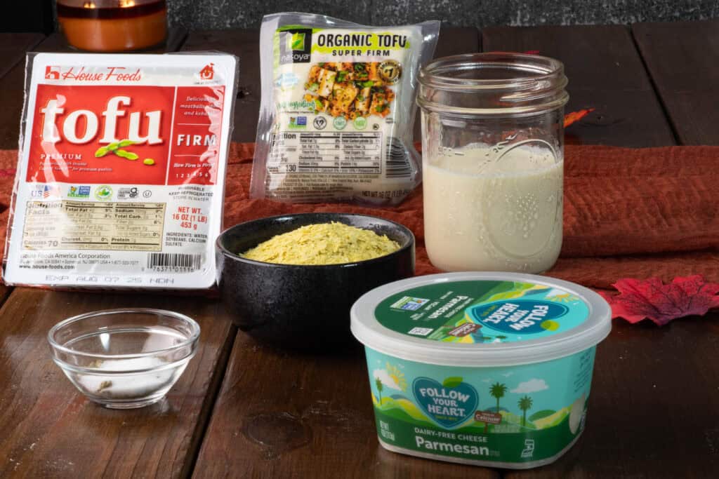 Ricotta ingredients sitting on a wooden table - firm tofu, super firm tofu, soy milk, nutritional yeast, vegan parmesan and spices.