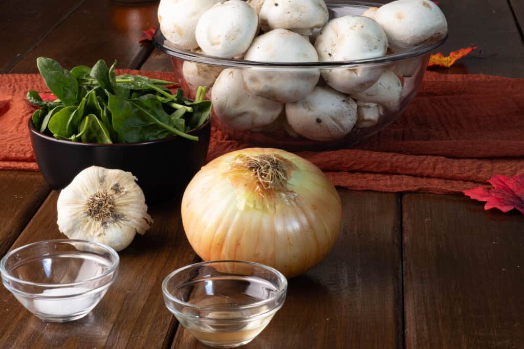 Spinach and mushroom lasagna layer ingredients on a wooden table - mushrooms, spinach, garlic, onion, vinegar and salt.