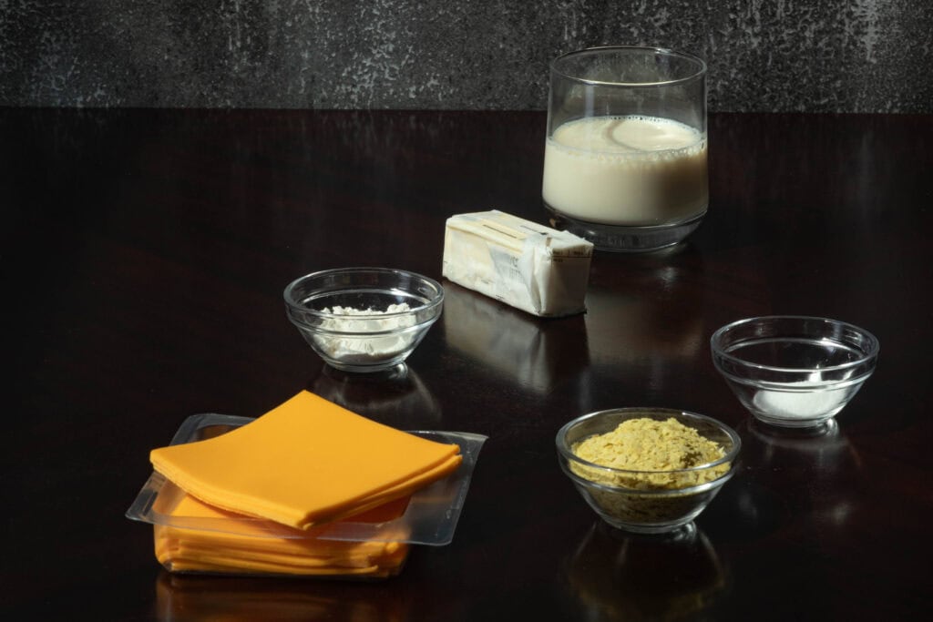 Vegan cheese sauce ingredients sitting on a wooden table - vegan cheese slices, flour, butter, soy milk, nutritional yeast and sugar.