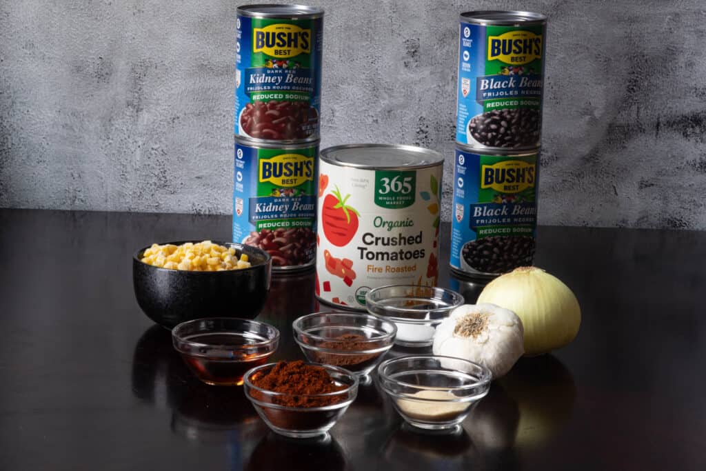 High Protein Vegan Chili ingredients sitting on a wooden table - kidney beans, black beans, crushed fire roasted tomatoes, corn, onion, garlic, syrup, cocoa powder, and various spices.