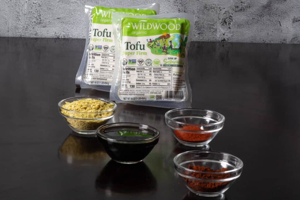 Tofu crumble ingredients sitting on a wooden table - super firm tofu, nutritional yeast, soy sauce, and spices.