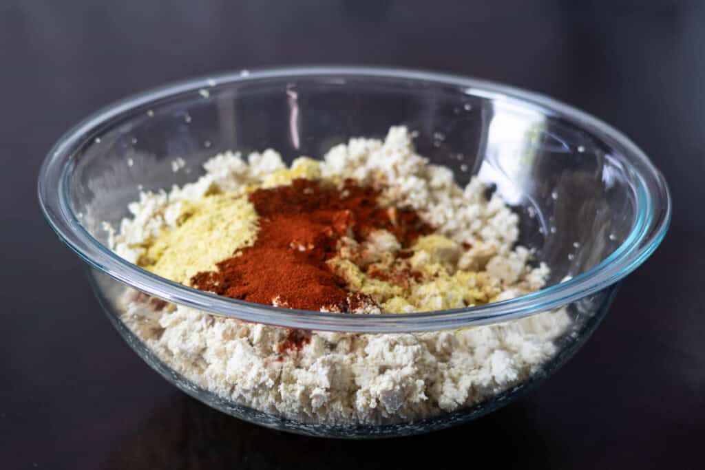 Large glass bowl with crumbled tofu, nutritional yeast, soy sauce and spices.