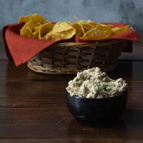 Bowl of vegan crab rangoon dip with basket of tortilla chips in the background.