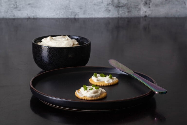 Small plate with two crackers topped with vegan cream cheese and scallions. Bowl with remaining cream cheese in the background.