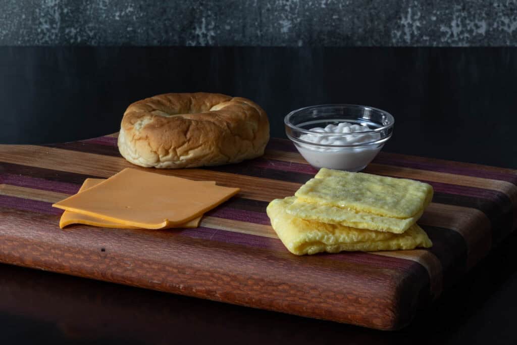 Breakfast sandwich ingredients on a wooden cutting board - bagel, vegan cheese slices, vegan egg patties, and mayo.