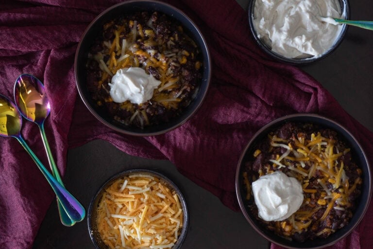Two bowls of chili topped with melted vegan cheese and sour cream, a smaller bowl of sour cream, a smaller bowl of shredded cheese and two spoons.