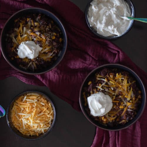 Two bowls of chili topped with melted vegan cheese and sour cream, a smaller bowl of sour cream, a smaller bowl of shredded cheese and two spoons.