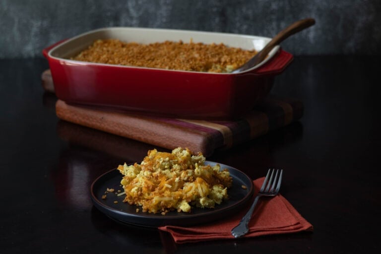 Plate of high protein vegan mac and cheese topped with crispy TVP breadcrumbs. Large casserole dish with remaining mac and cheese in the background.