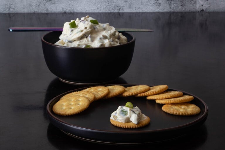Plate of crackers, with one cracker topped with holiday dip. Bowl of holiday cream cheese dip in the background.