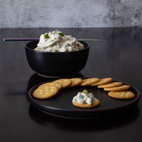 Plate of crackers, with one cracker topped with holiday dip. Bowl of holiday cream cheese dip in the background.