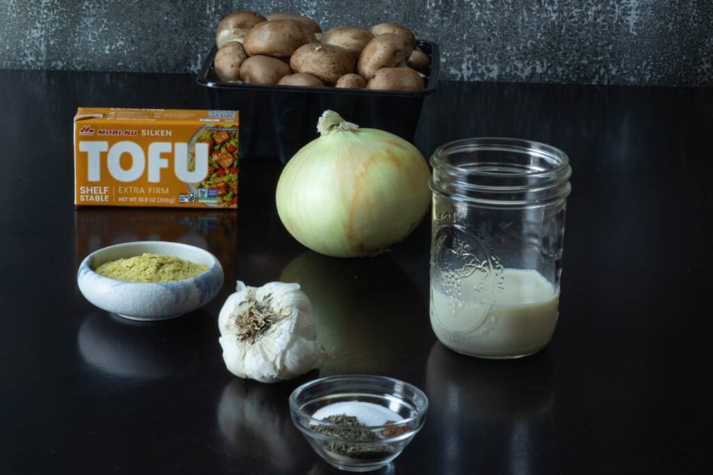 Vegan Mushroom Soup ingredients sitting on a table - silken tofu, mushrooms, nutritional yeast, onion, garlic, soy. milk and spices.