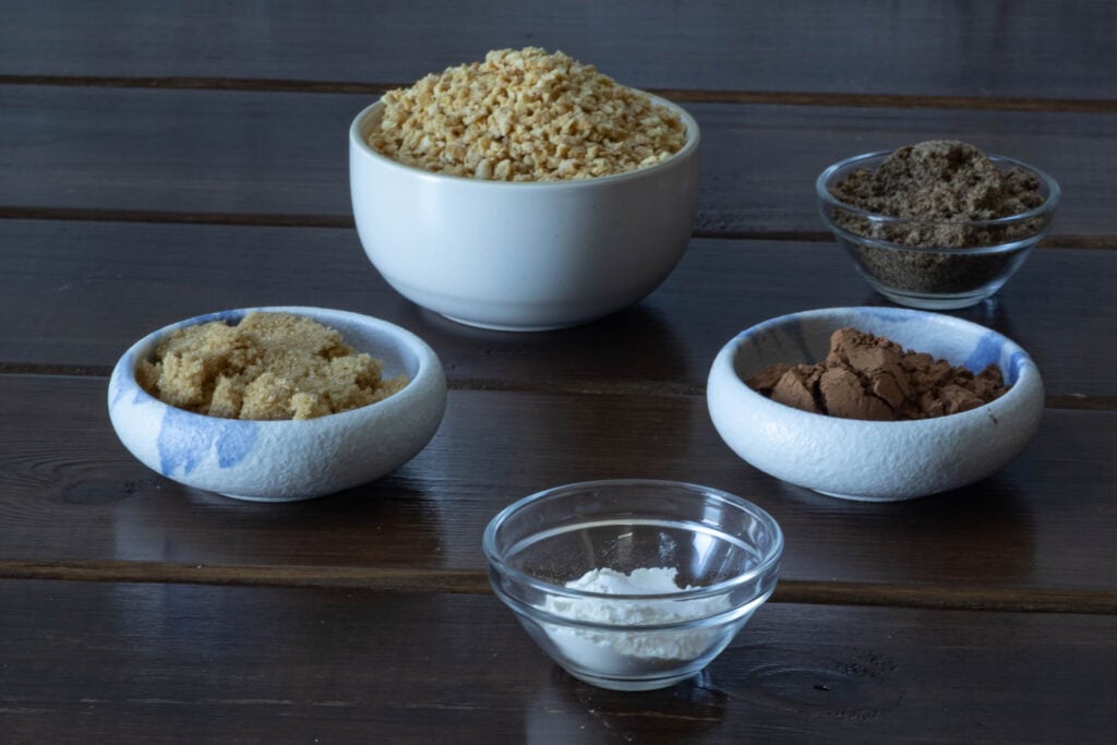 Ingredients for chocolate TVP cheesecake crust sitting on a wooden table - brown sugar, TVP, flour, cocoa powder, ground flax.