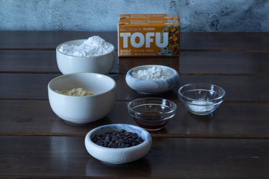 Peanut butter chocolate chip cheesecake ingredients sitting on a wooden table - powdered sugar, silken tofu, peanut butter powder, flour, vanilla extract, salt, baking powder, and mini chocolate chips.