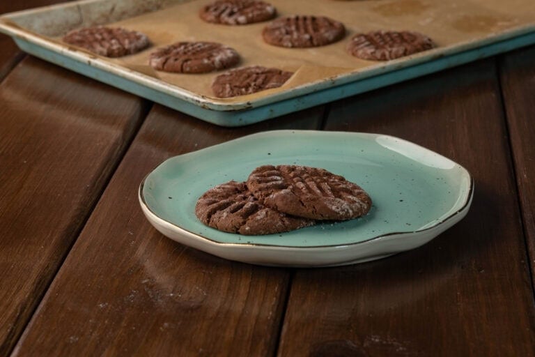 Two peanut butter chocolate cookies sitting on a plate with a baking sheet with the remaining cookies in the background.