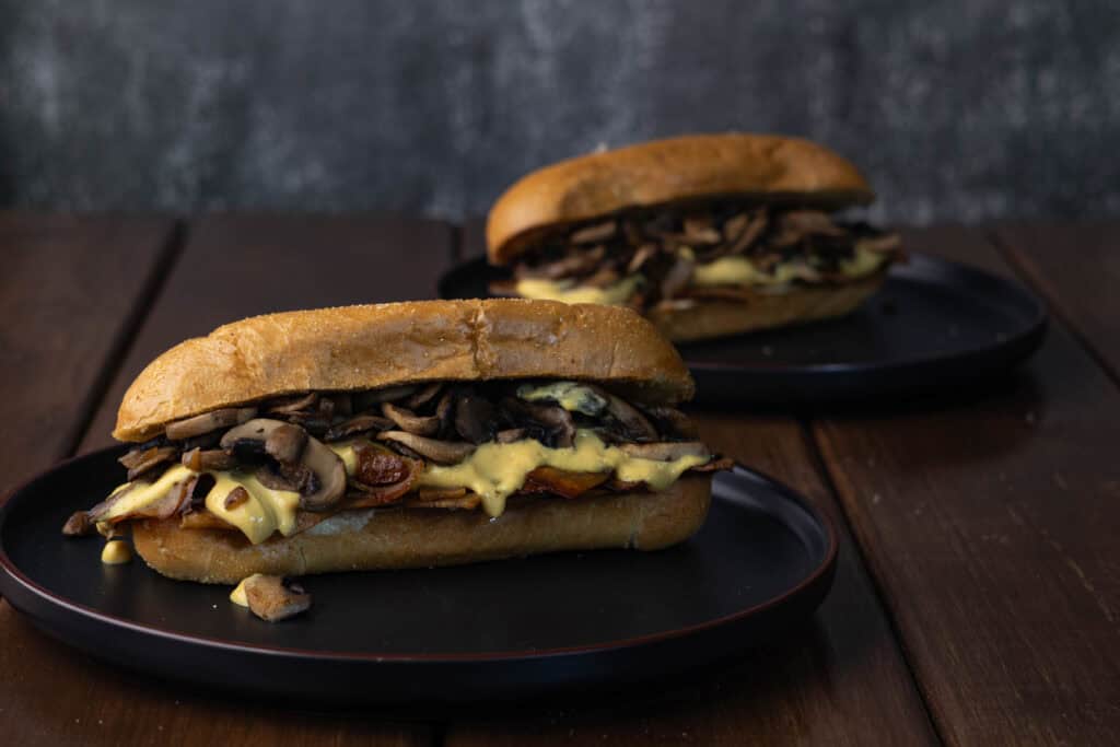 Two vegan Philly Cheesesteak sandwiches with thinly sliced and marinaded tofu, sauteed onions and mushrooms and a vegan cheese sauce.