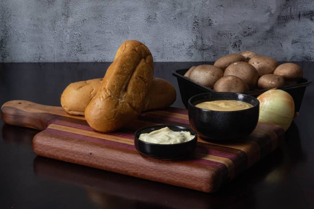 Vegan philly cheese steak ingredients sitting on a wooden cutting board - hoagie buns, mayo, cheese sauce, mushrooms, and an onion.