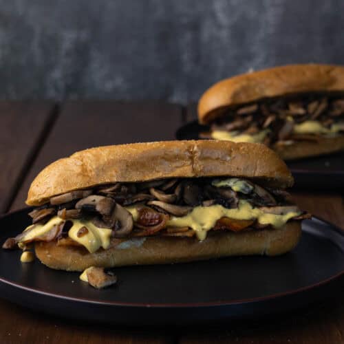 Two vegan Philly Cheesesteak sandwiches with thinly sliced and marinaded tofu, sauteed onions and mushrooms and a vegan cheese sauce.